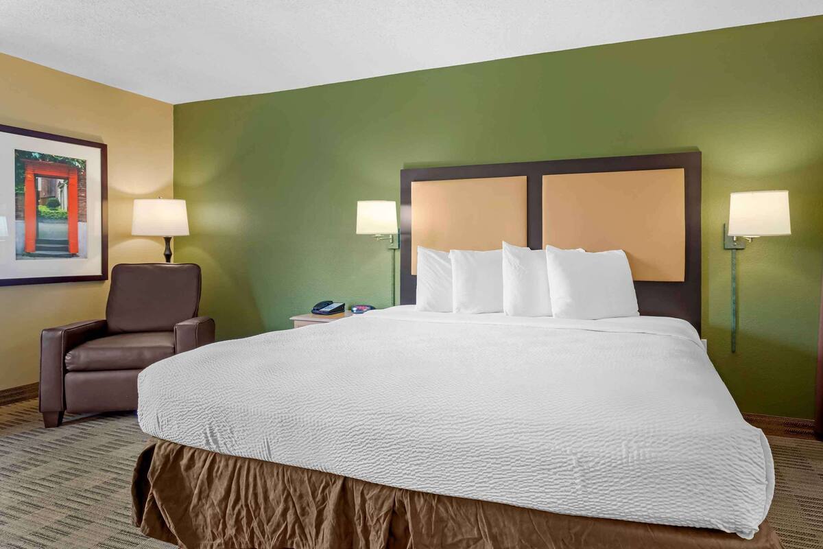Una habitació d'hotel amb un llit gran, dues làmpades de nit, una cadira de cuir marró i un quadre encarregat a la paret.