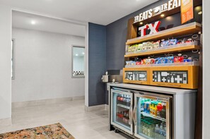 Un bar de aperitivos con una variedad de productos envaselados, bebidas y un refrigerador, además de un cartel que indica su ubicación.