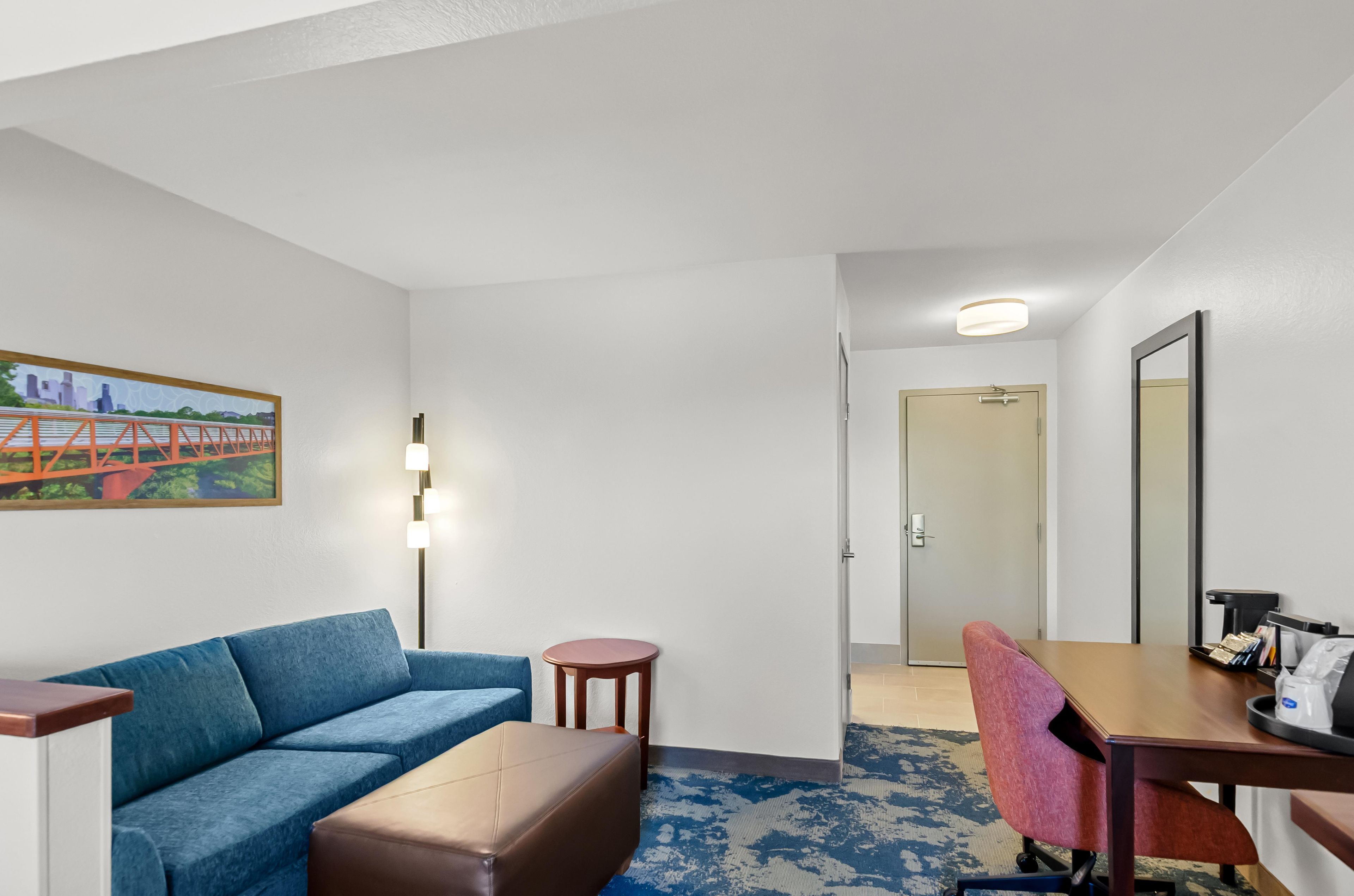 A hotel room with a blue sofa, a small round table, a wooden desk, and a chair. There is a painting on the wall and a door leading to another room.