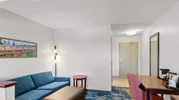 A hotel room with a blue sofa, a small round table, a wooden desk, and a chair. There is a painting on the wall and a door leading to another room.