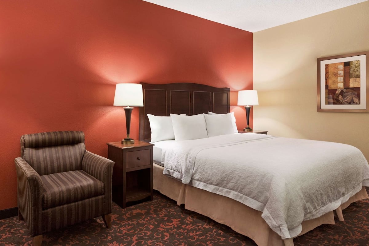 A hotel room with a bed, two bedside lamps, a chair, a nightstand, and a framed picture on the wall.