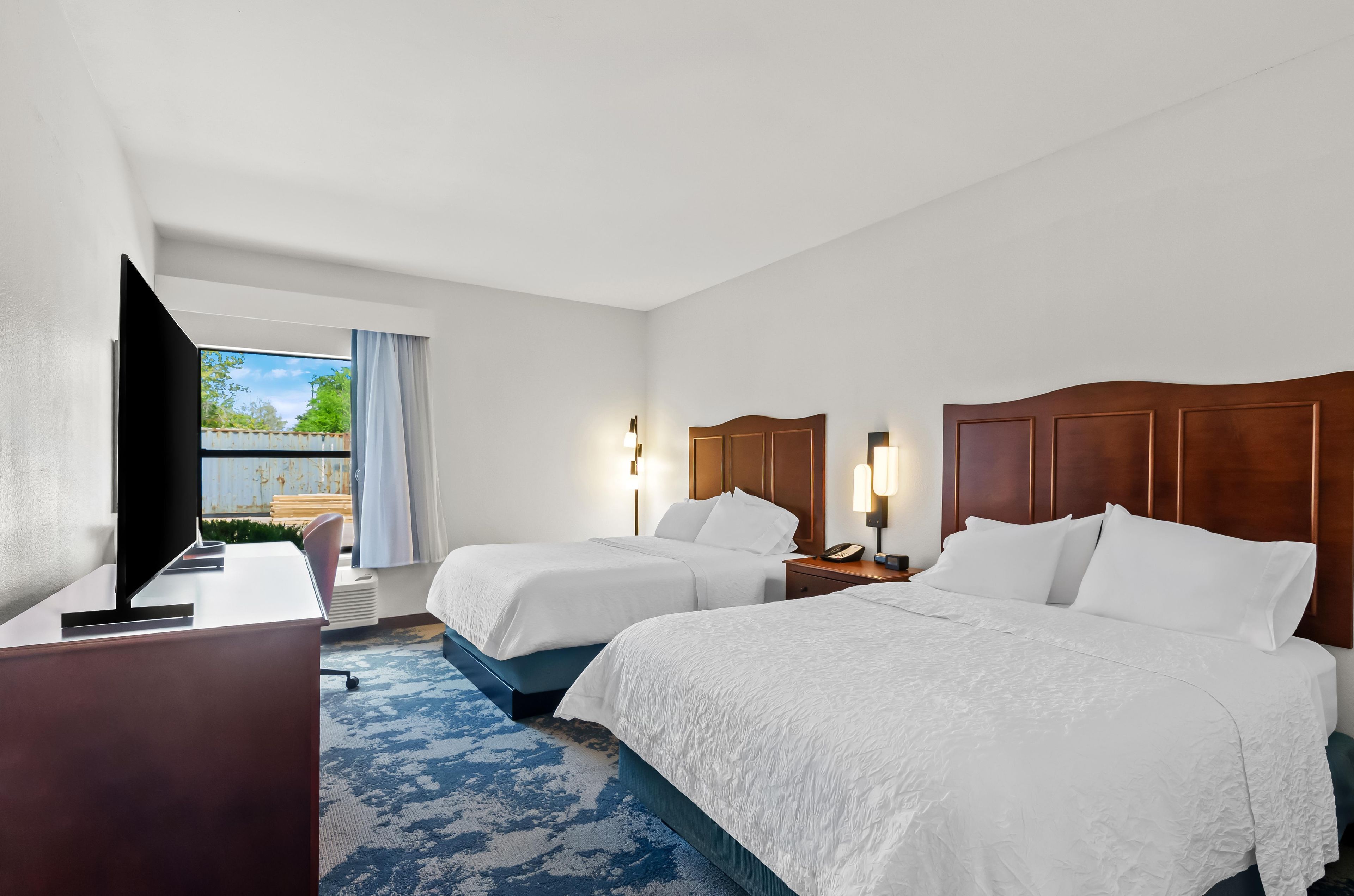 A hotel room with two beds, a desk, a television, and a window with curtains.