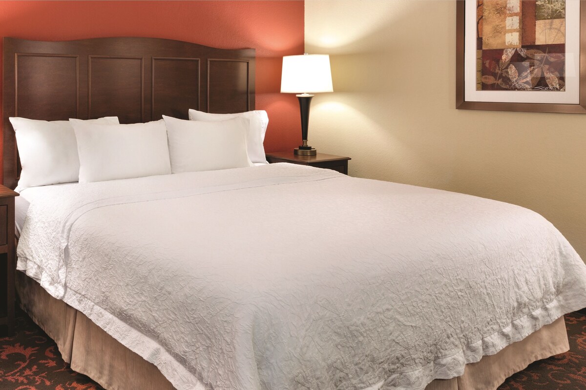 A neatly made bed with white linens and a wooden headboard, a bedside lamp, and a framed artwork on the wall.