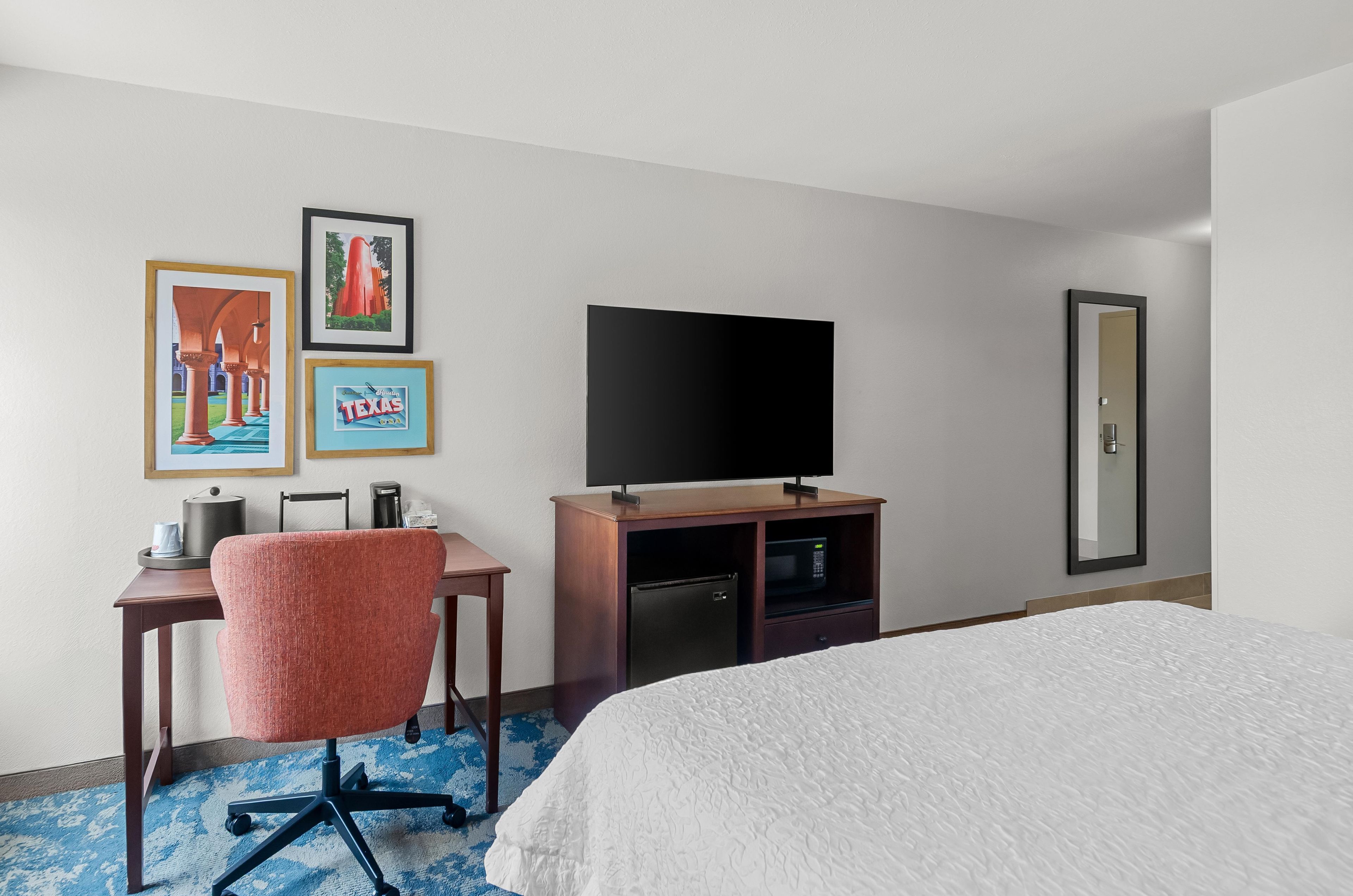 A hotel room with a bed, a desk with a chair, a flat-screen TV, a microwave, and framed pictures on the wall.
