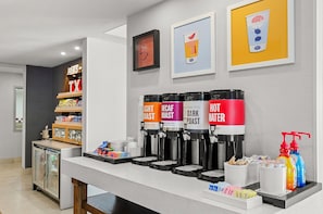 Estação de café com diversos dispensadores de bebidas, um refrigerador de exposição e um balcão com lanches e acompanhamentos.