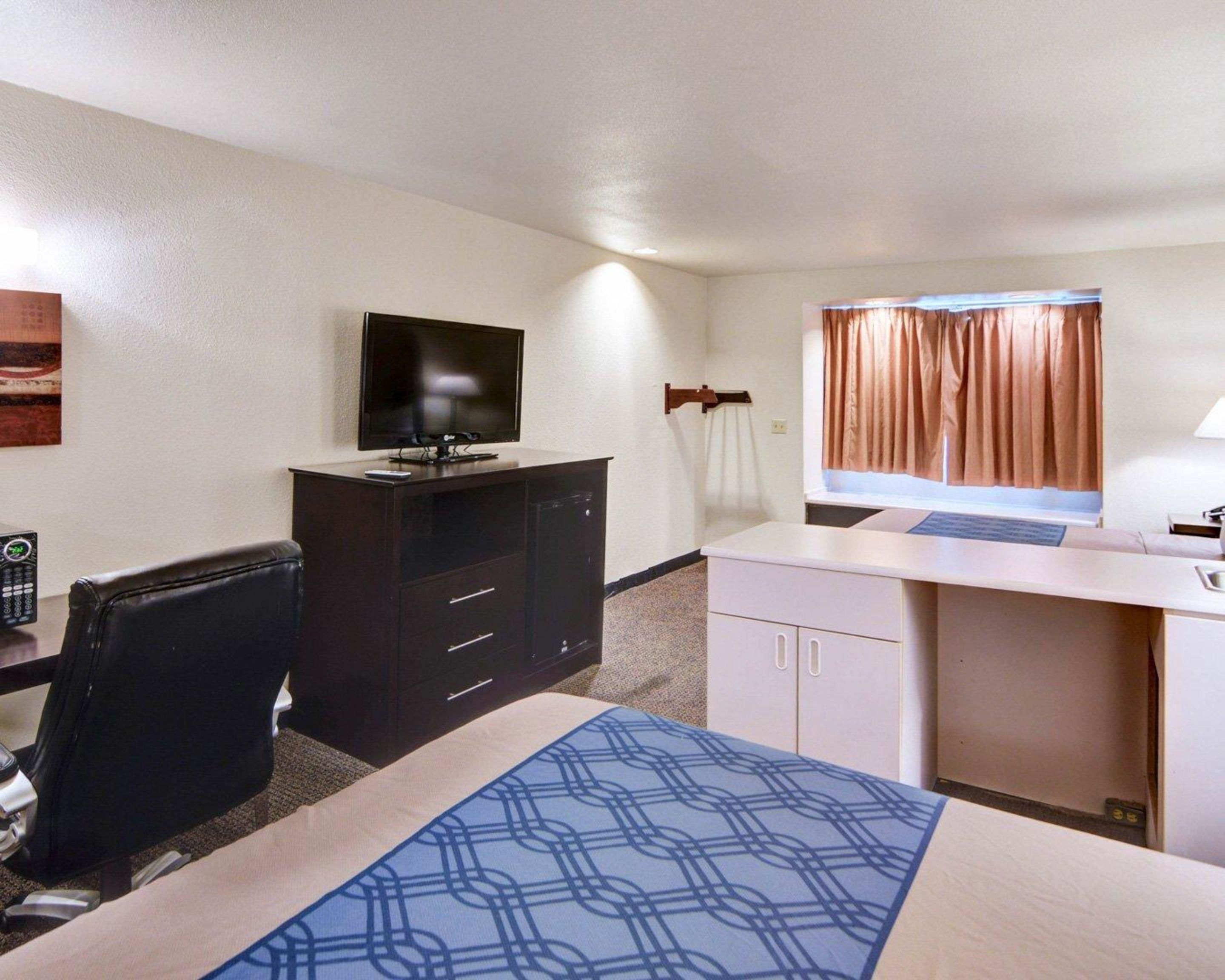A hotel room with a bed, a desk, a chair, a television, a window with curtains, and a wall-mounted lamp.
