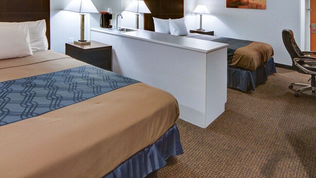 Quarto de hotel com uma cama grande, uma cama menor, uma escrivaninha e uma cadeira. Há duas luminárias na escrivaninha e um quadro na parede.