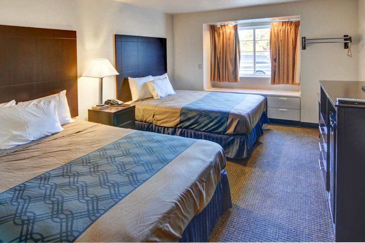 A hotel room with two beds, a desk, a lamp, a window with curtains, and a dresser.