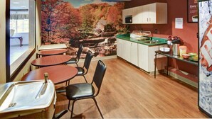 Uma sala de jantar com um mural de paisagem, mesas e cadeiras de madeira, área de cozinha com pia e armários, e uma geladeira.