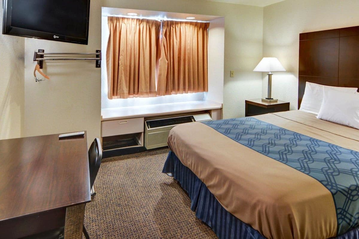 A hotel room with a bed, a desk, a television mounted on the wall, a window with curtains, a bedside lamp, and a headboard.