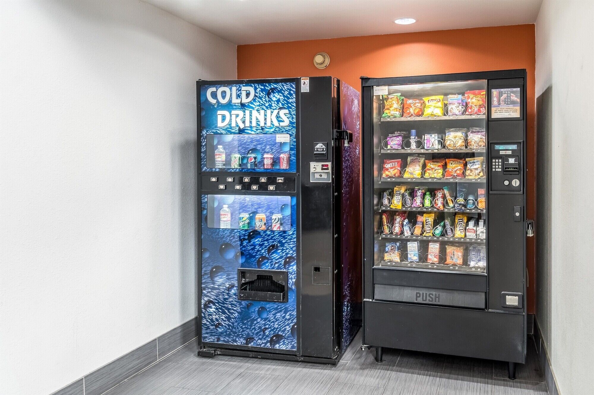 Dos máquinas expendedoras, una con la etiqueta 'COLD DRINKS' y la otra 'PUSH', en un pasillo con piso de baldosas y paredes anaranjadas.
