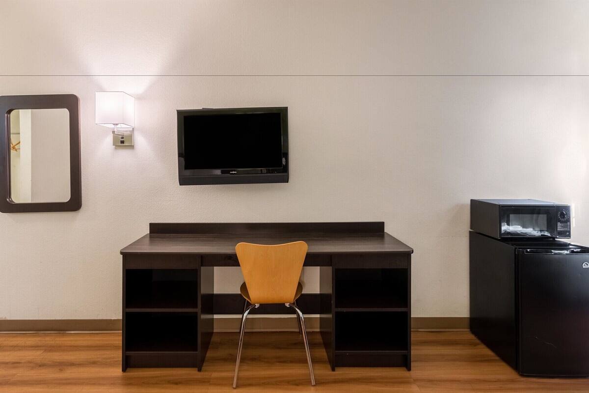 Une pièce avec un bureau en bois foncé, une chaise, un téléviseur à écran plat fixé au mur et un réfrigérateur noir.