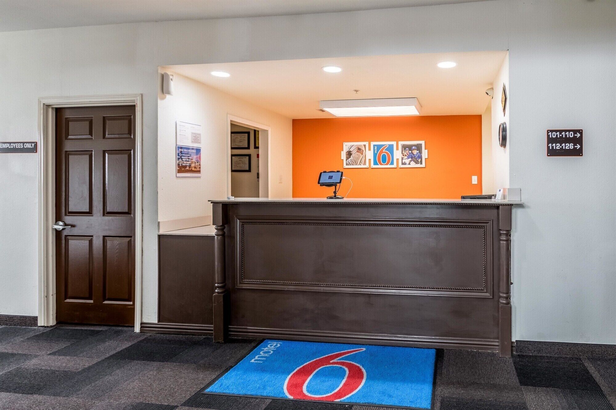 A reception desk with a digital screen, a blue mat with a red number six, and directional signs for room numbers.