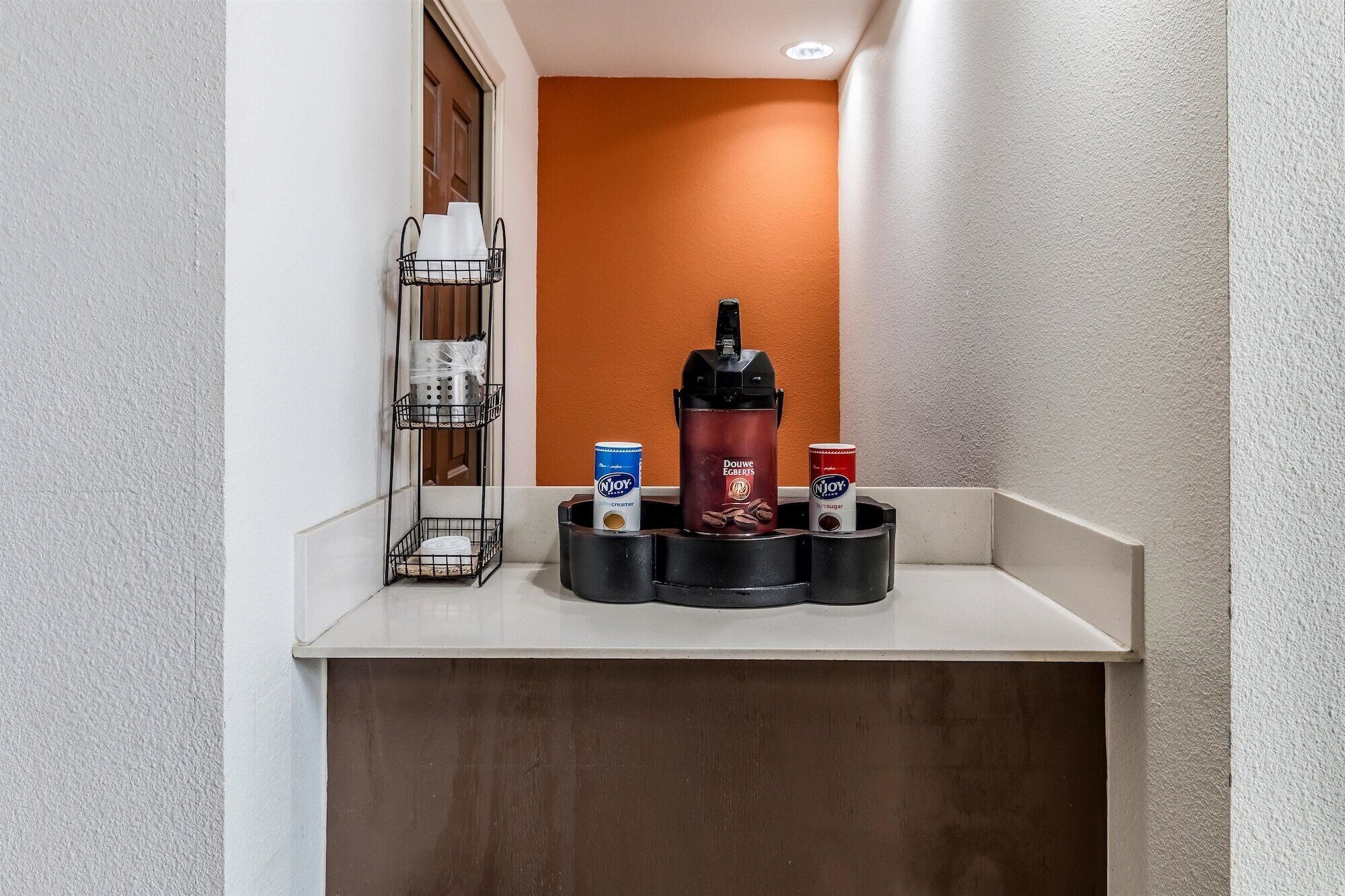 Un coin café avec une machine à café et deux boîtes de Njoy, une contenant du sucre et l’autre de la crème, sur un comptoir blanc.