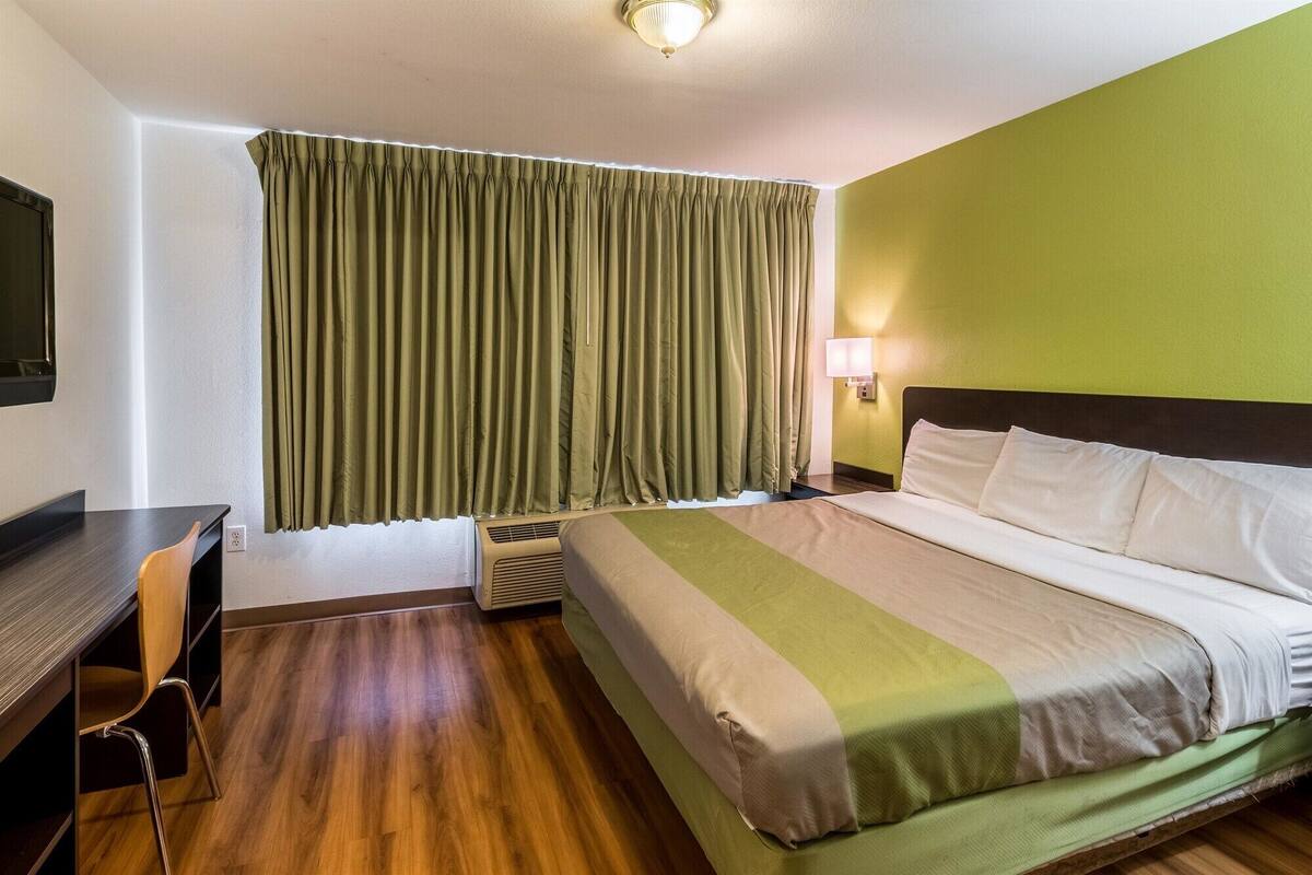 Une chambre d’hôtel avec un lit, un bureau, une chaise, une lampe et une fenêtre avec des rideaux.