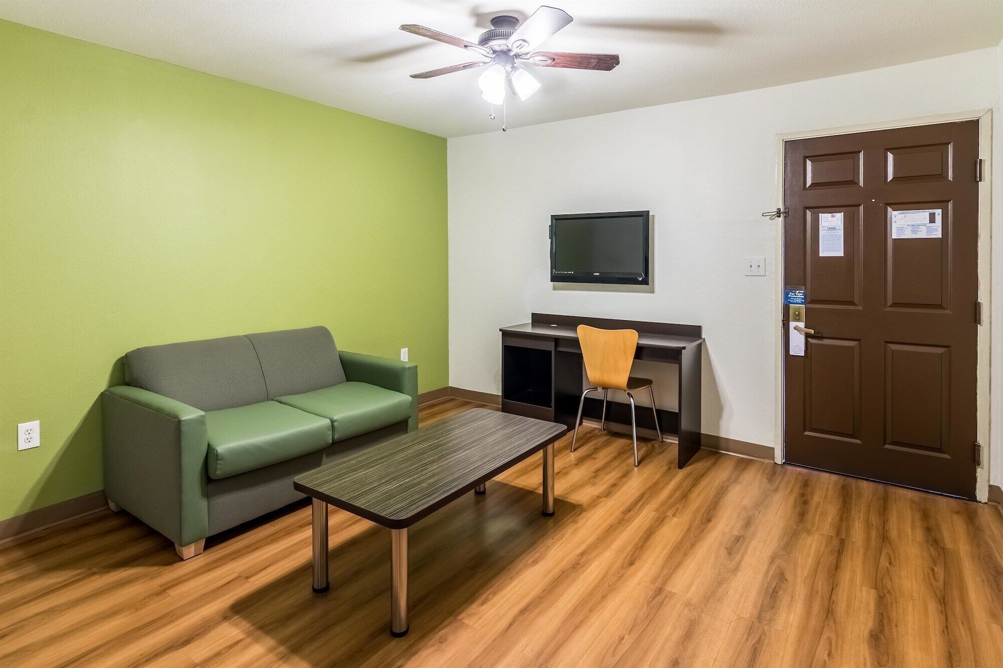 Une pièce avec un mur vert, un plancher en bois, un canapé vert, une petite table, une chaise, un téléviseur fixé au mur et une porte avec une serrure.
