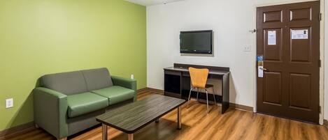 A room with a green wall, a wooden floor, a green sofa, a small table, a chair, a TV mounted on the wall, and a door with a lock.