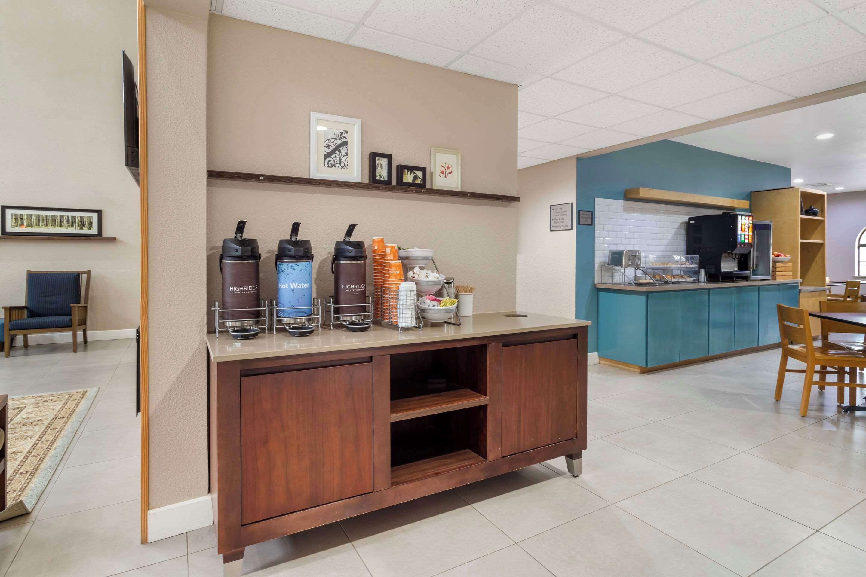 Zona de recepción de un hotel con mostrador de madera, dispensadores de agua caliente y una máquina de café. En la pared hay cuadros enmarcados, un televisor y un mostrador azul al fondo.