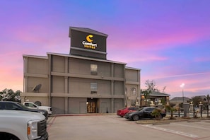 A Comfort Suites hotel with a prominent sign, a parking lot with several cars, and a clear sky.