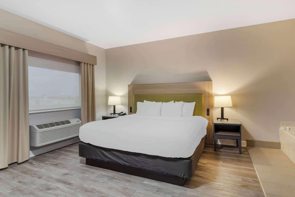 Une chambre d’hôtel avec un grand lit, deux tables de chevet avec des lampes, une fenêtre avec des rideaux, un appareil de climatisation et une petite salle de bain.