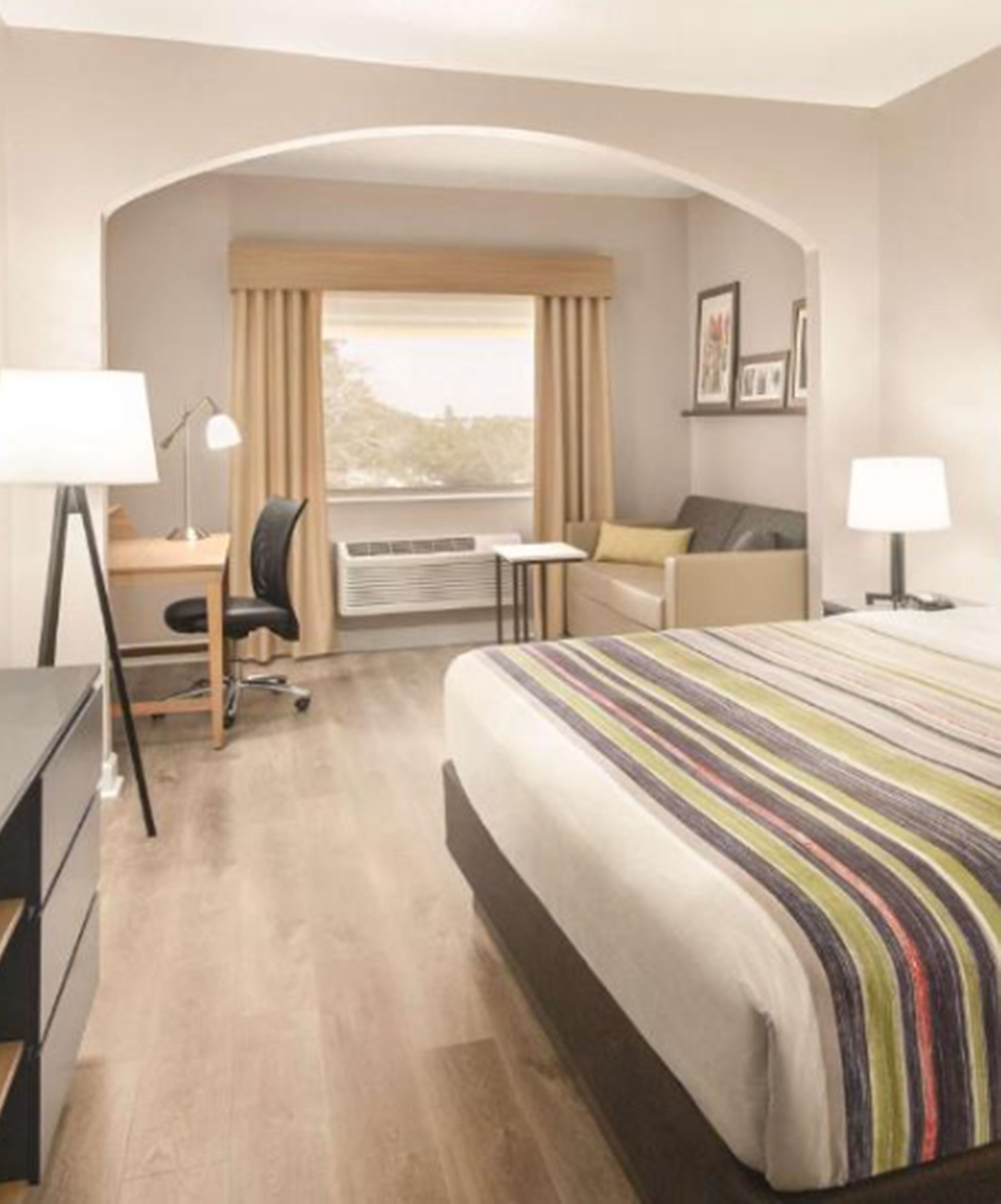 A hotel room with a bed, desk, chair, and an air conditioning unit.