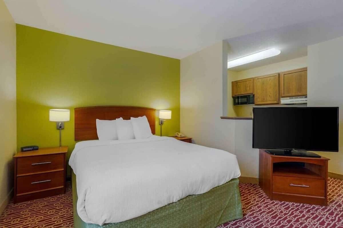 A hotel room with a bed, bedside lamps, a television, a nightstand, and a microwave in the kitchen area.