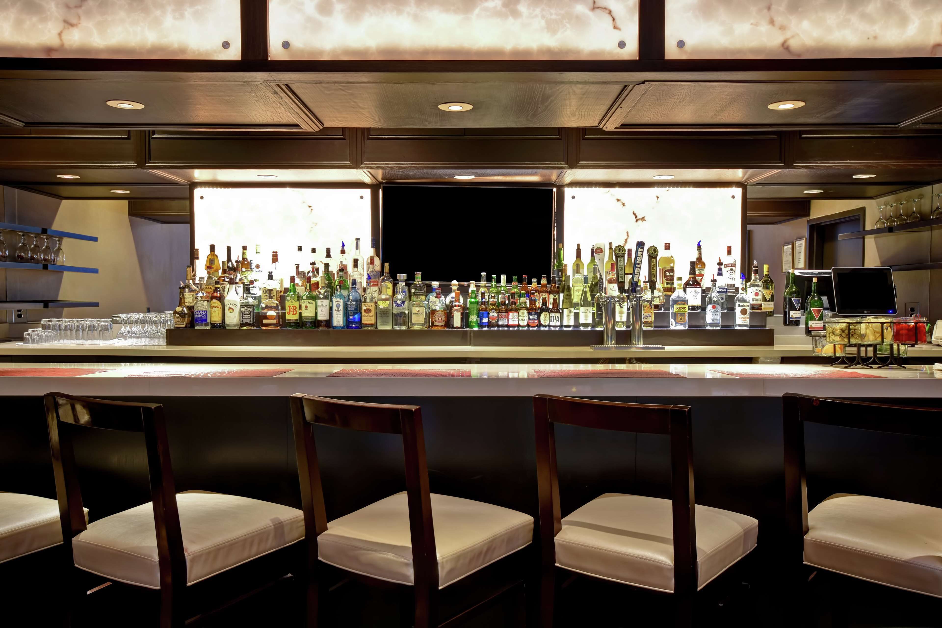 A bar area with a variety of liquor bottles, glasses, and a laptop on the counter. There are four wooden bar chairs in front.