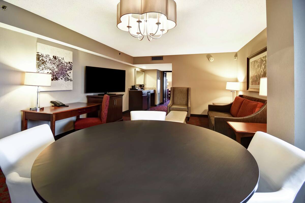 Une chambre d’hôtel avec une table de salle à manger ronde, des chaises blanches, un téléviseur à écran plat, un bureau avec un téléphone et un canapé agrémenté d’un coussin rouge.