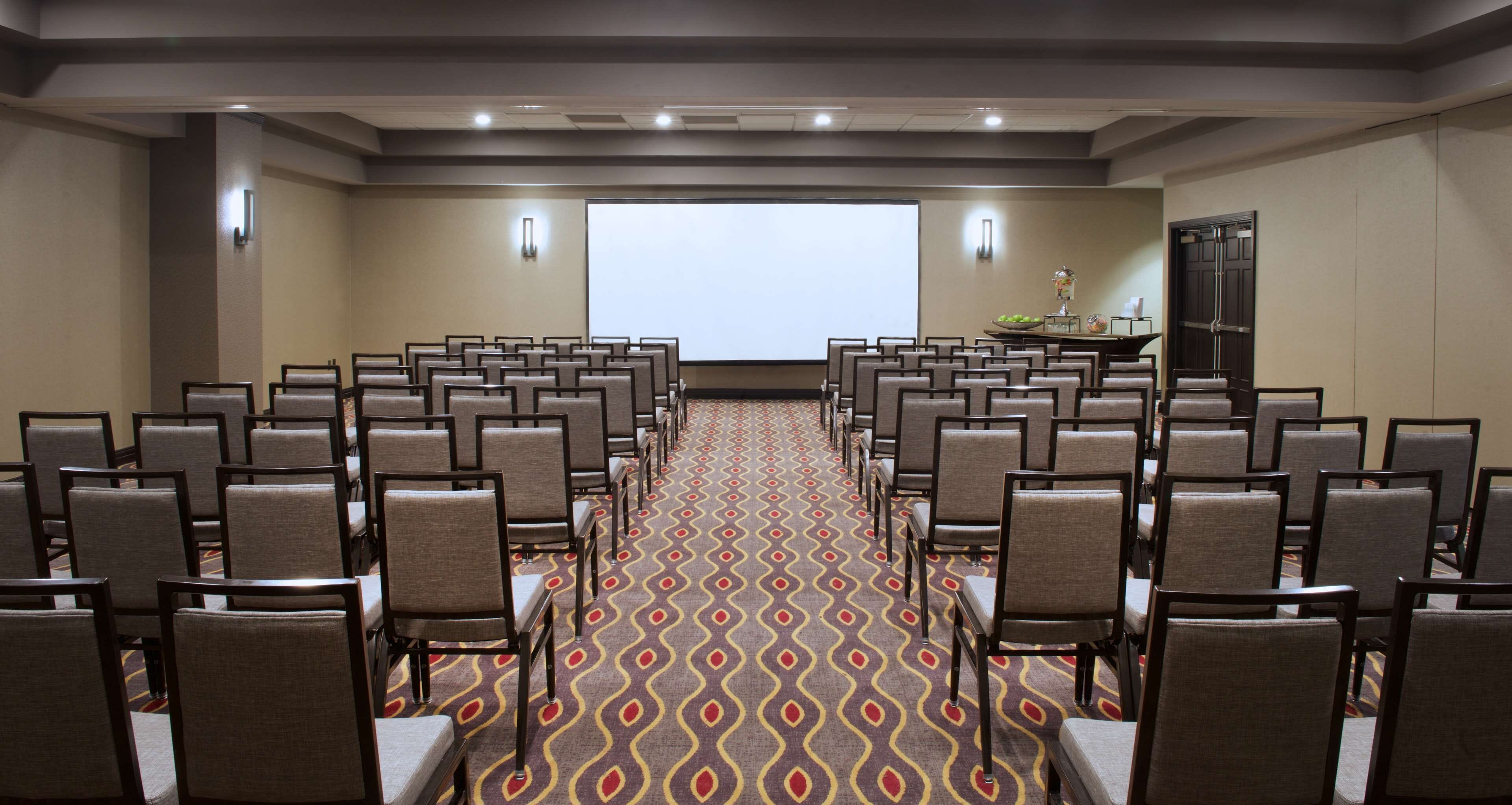 A conference room with rows of chairs facing a large screen, a patterned carpet, and a small table with a bowl of fruit.