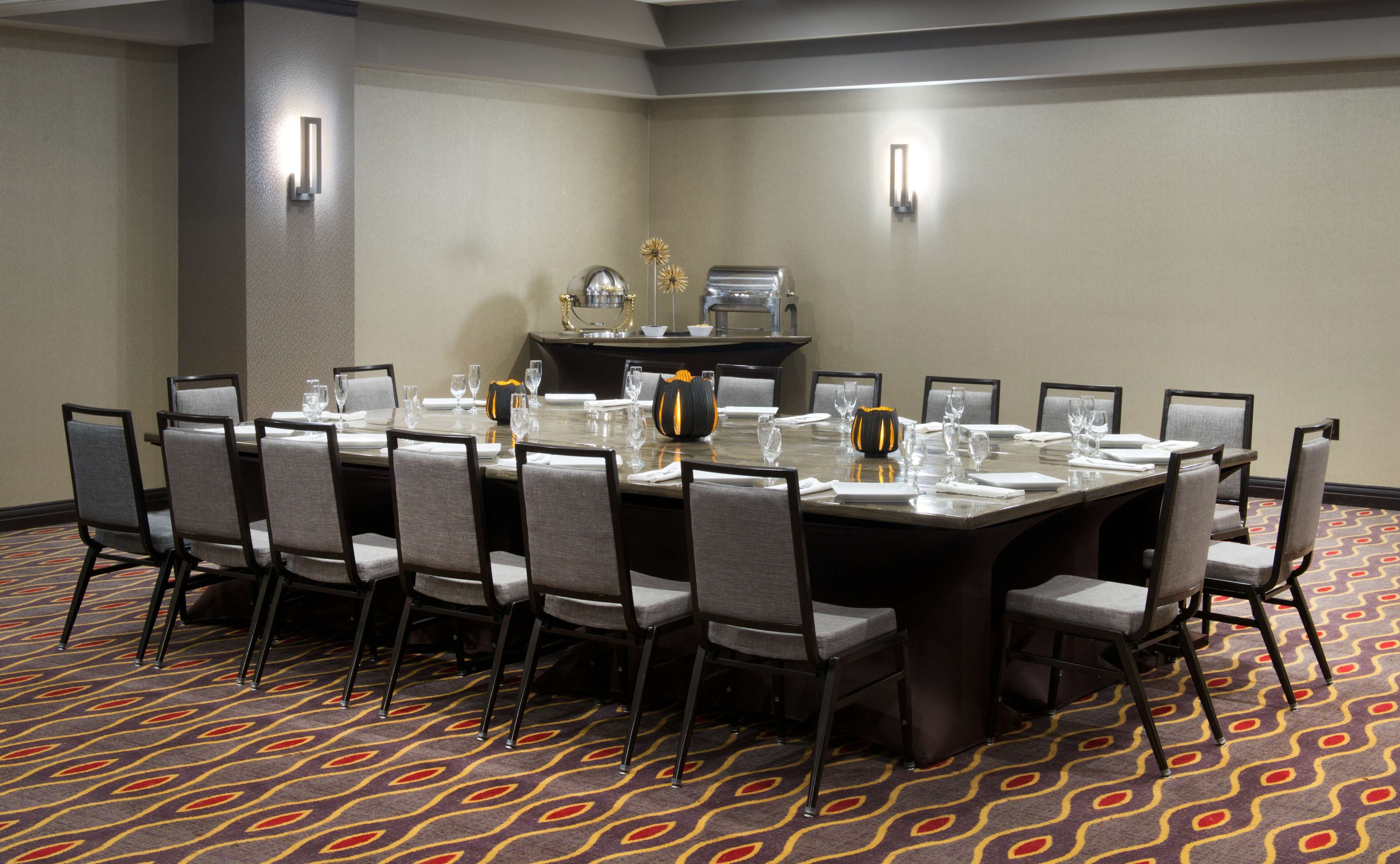 A conference room with a large rectangular table, grey upholstered chairs, and a patterned carpet.