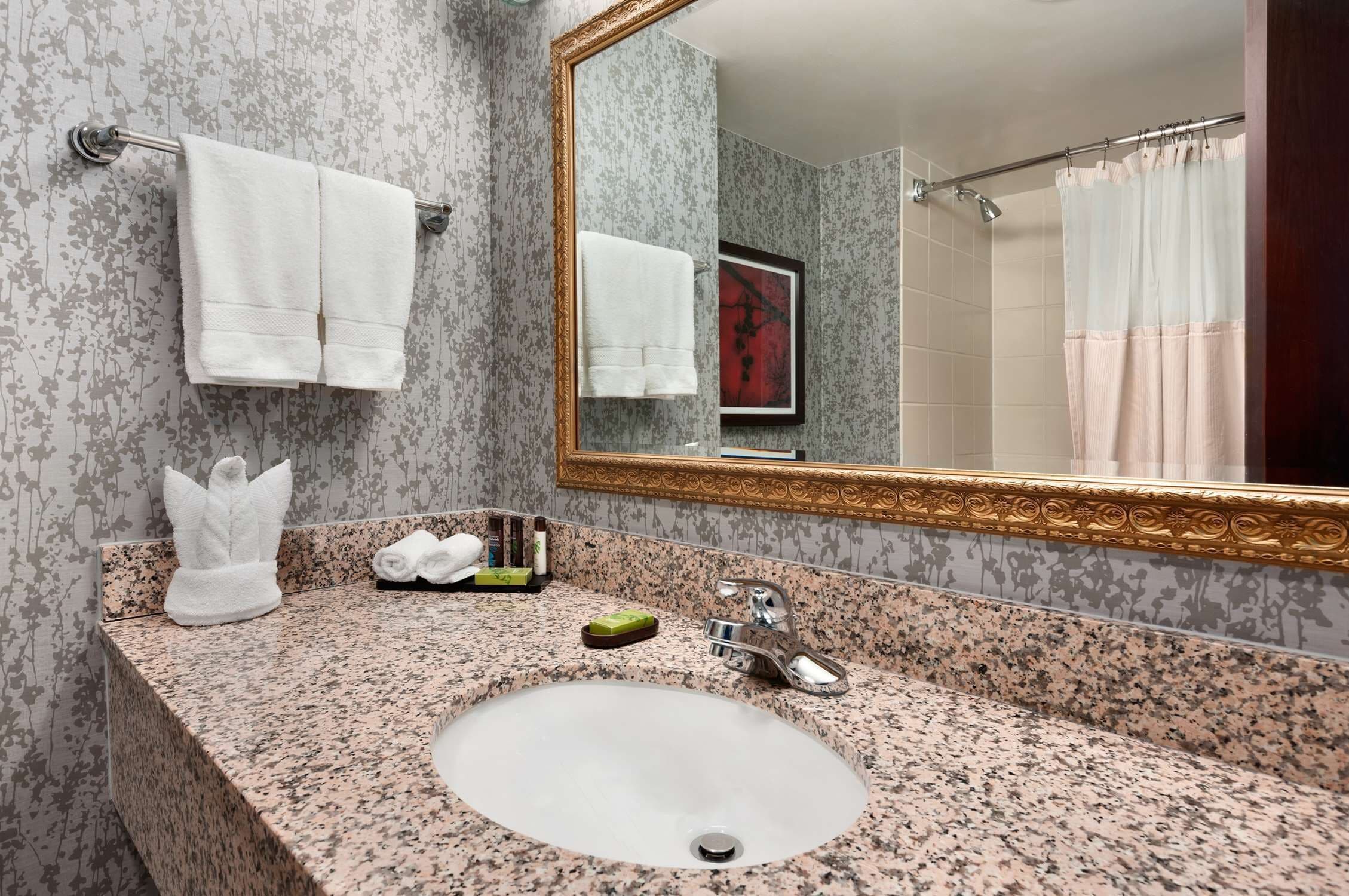 Un baño con lavamanos de encimera de granito, dos toallas blancas, un espejo grande y una cortina de ducha.