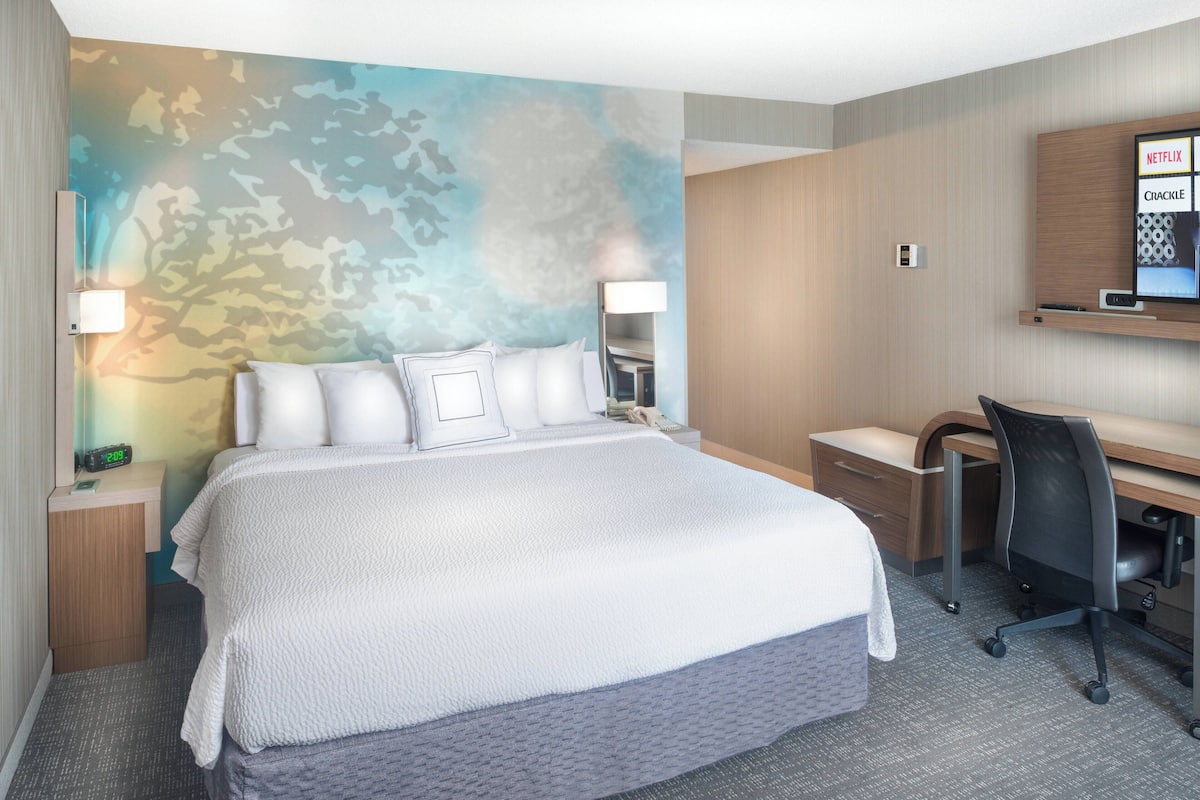 Ein Hotelzimmer mit einem großen Bett, einem Schreibtisch, einem Sessel, einem Fernseher und einer Wand mit einer Baummalerei.
