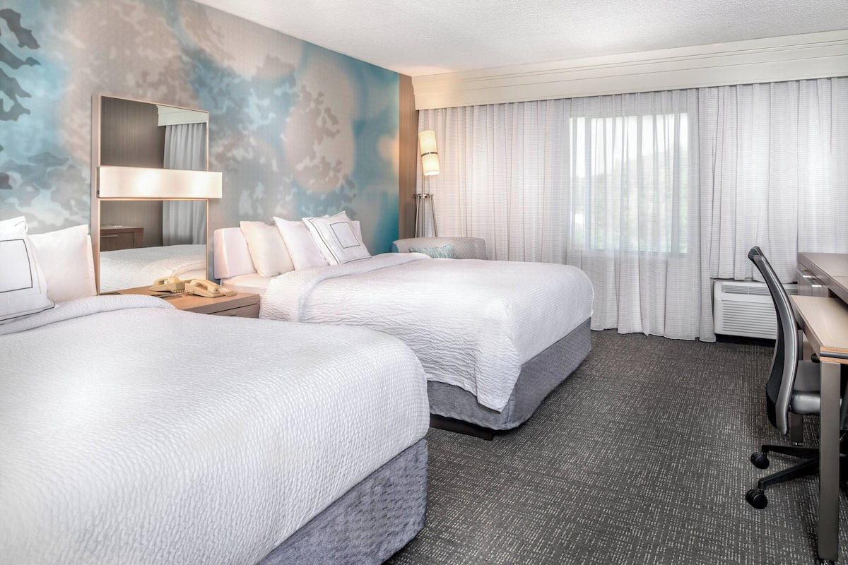 Ein Hotelzimmer mit zwei Betten, einem Schreibtisch, einem Stuhl, einer Lampe und einem großen Fenster mit Vorhängen.