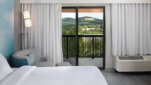 Ein Hotelzimmer mit einem großen Bett, einer Couch, einer Stehlampe, einem Heizkörper und einem Balkon mit Blick auf einen bewaldeten Hügel.