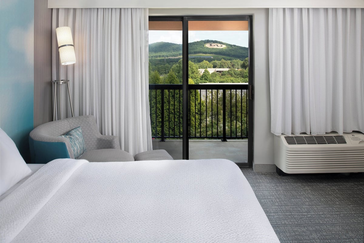 Ein Hotelzimmer mit einem großen Bett, einer Couch, einer Stehlampe, einem Heizkörper und einem Balkon mit Blick auf einen bewaldeten Hügel.