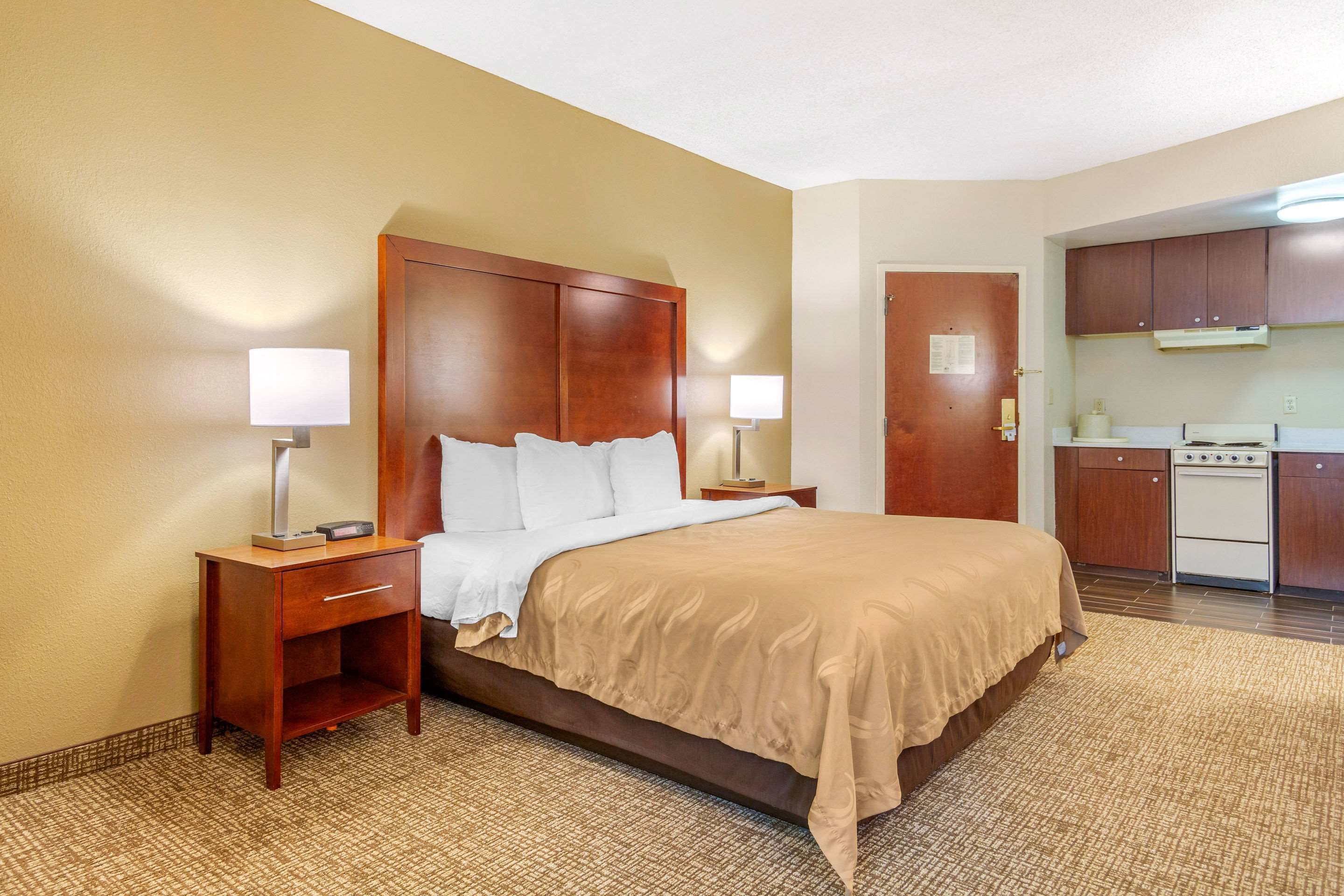 A hotel room with a large bed, bedside tables with lamps, a small desk, a kitchenette, and a door leading to another room.