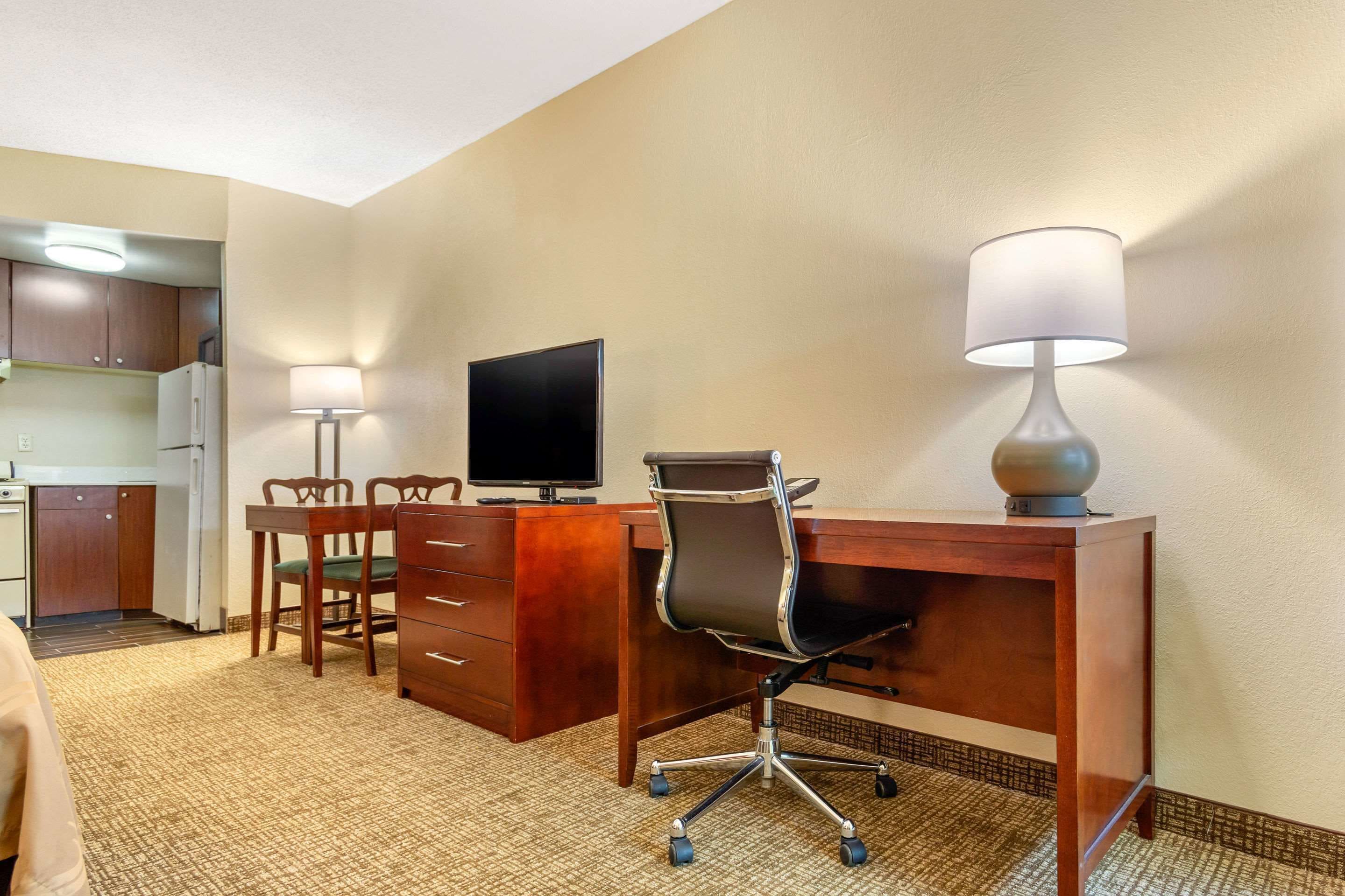 A hotel room with a wooden desk, a computer, a lamp, a chair, and a kitchenette in the background.