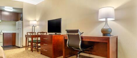 A hotel room with a wooden desk, a computer, a lamp, a chair, and a kitchenette in the background.