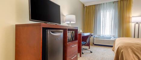 A hotel room with a flat-screen TV on a wooden dresser, a desk with a chair, a bed with a beige bedspread, and a window with yellow curtains.