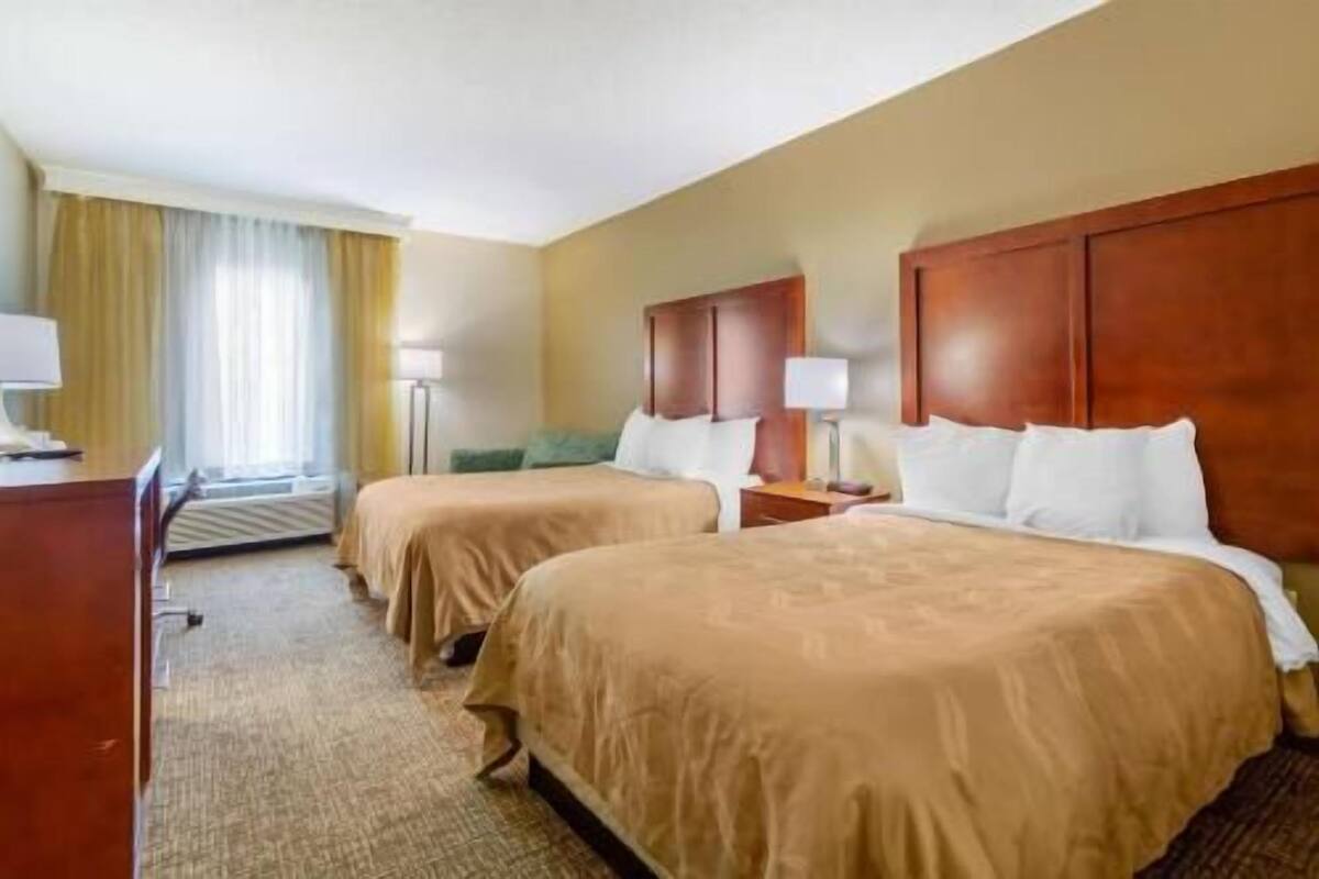 A hotel room with two beds, a desk, a chair, a lamp, and a window with curtains.