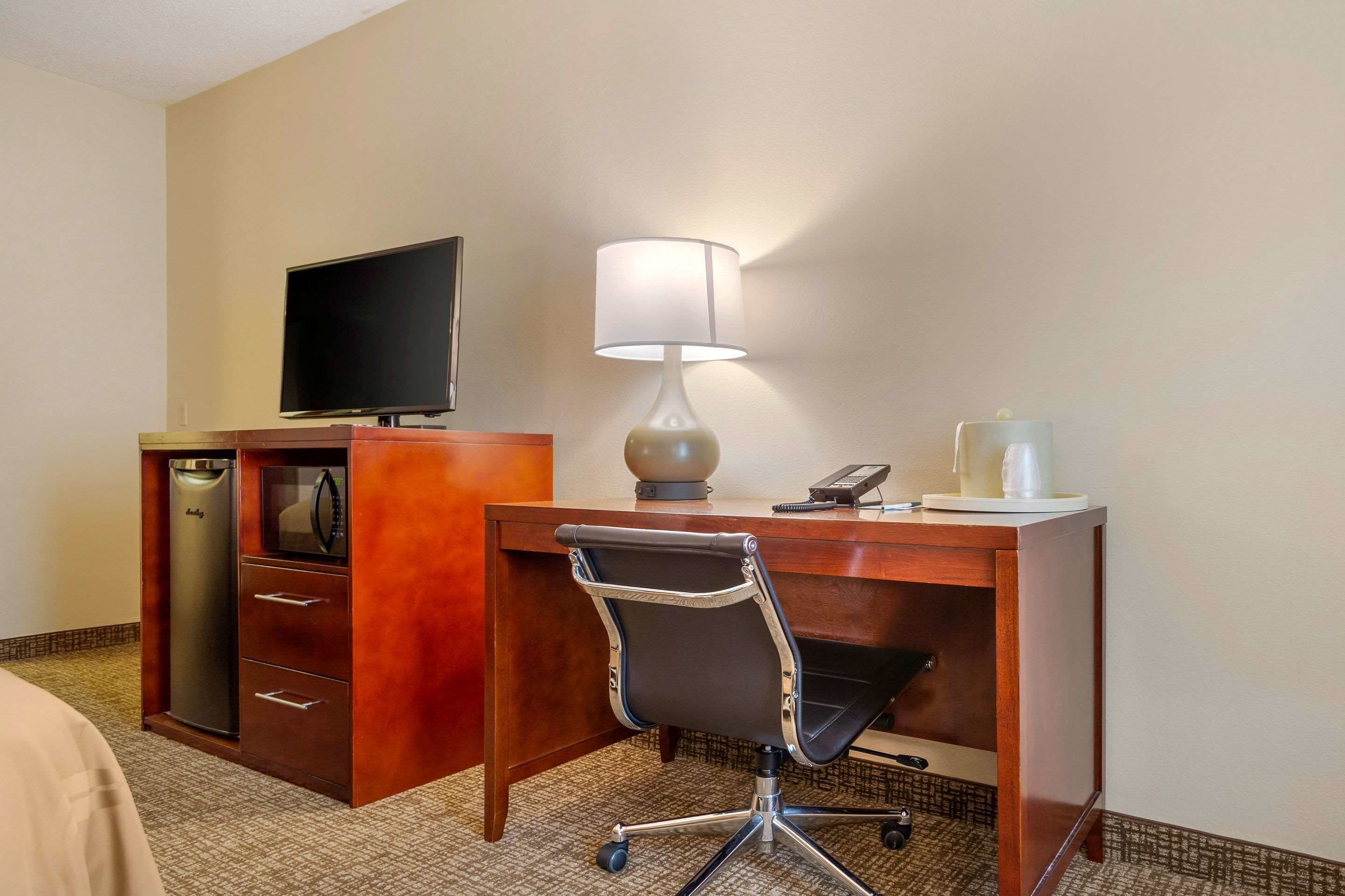 Habitación de hotel con un escritorio de madera, una silla de oficina negra, un televisor, una lámpara, un microondas y un minibar.