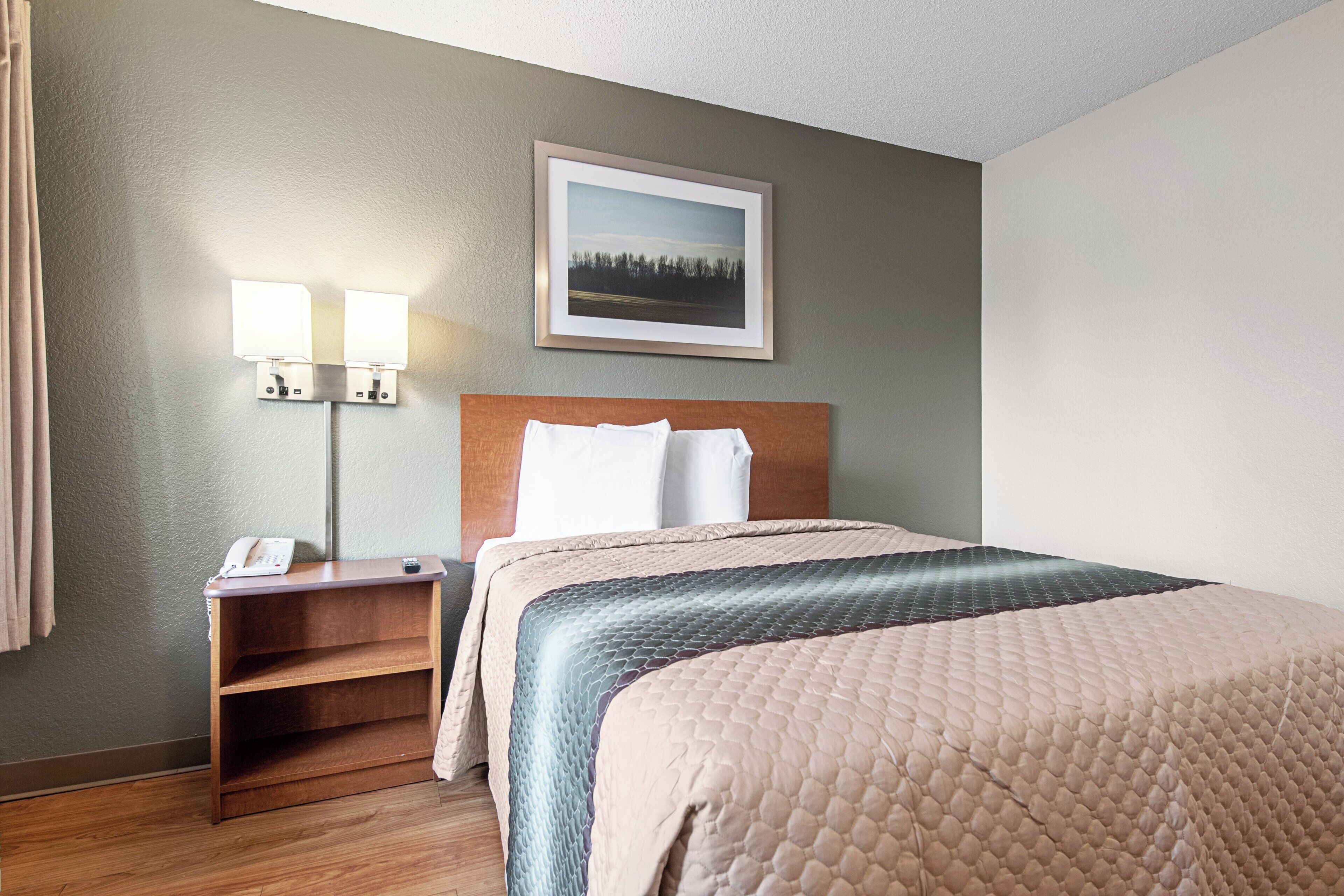 A hotel room with a bed, bedside lamps, a nightstand with a phone, a framed picture on the wall, and a wooden floor.