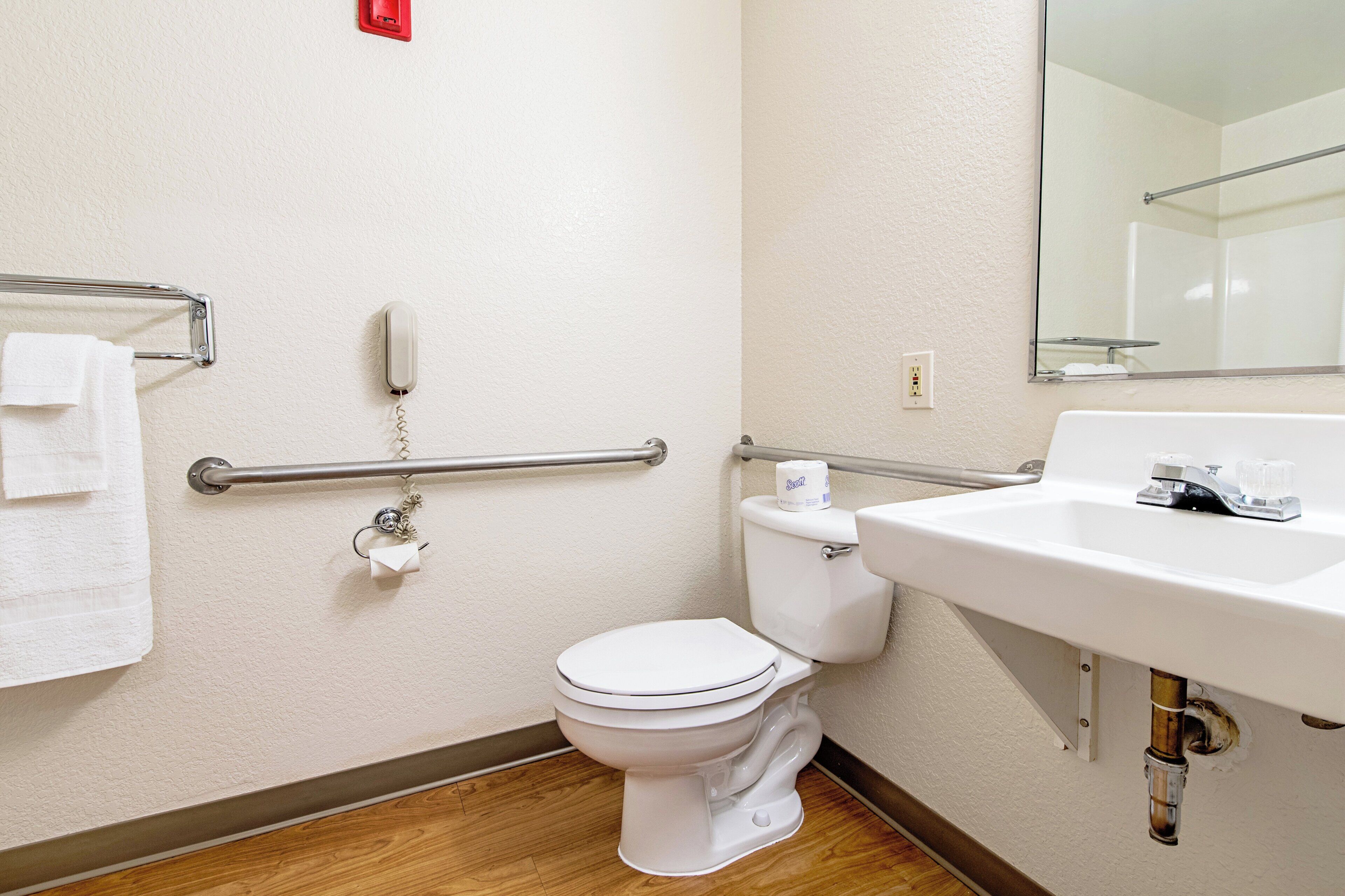 Un baño con inodoro blanco, lavabo, espejo, toallero y barra de apoyo.