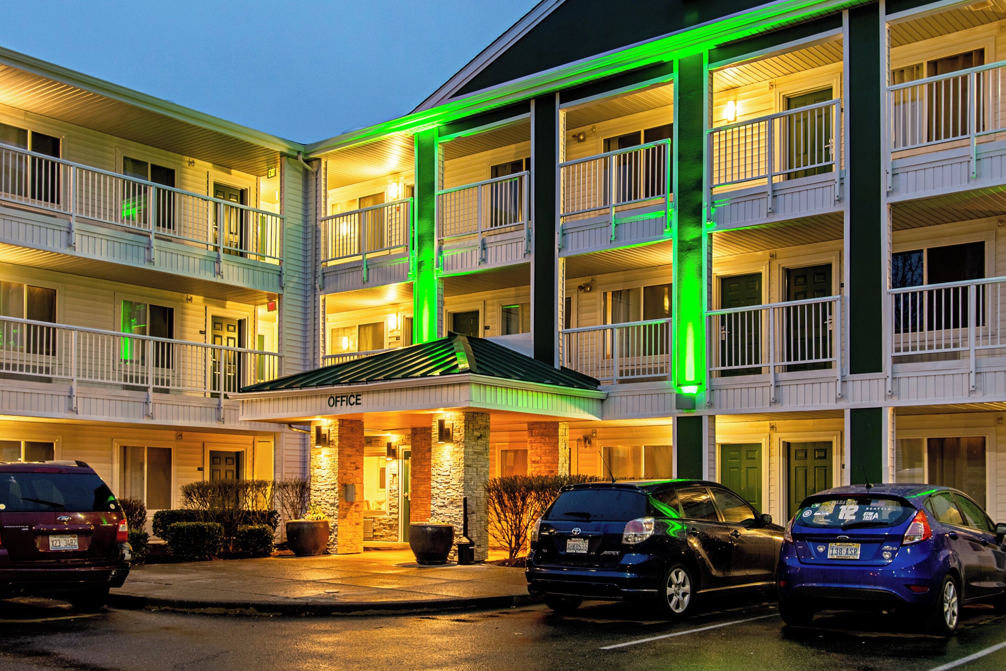A multi-story hotel with a central entrance, multiple balconies, and a parking area with cars.