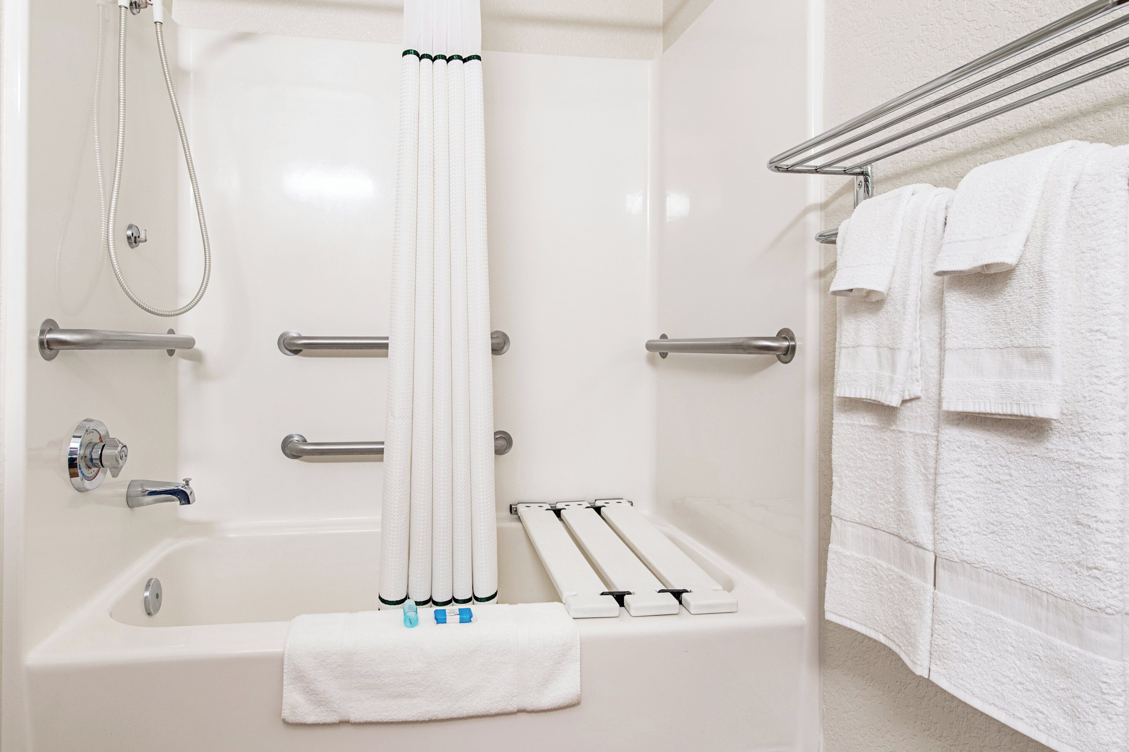 A bathroom with a bathtub, shower, handrails, a towel rack, and a towel placed on the floor.