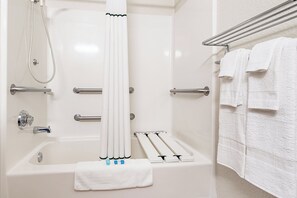 Ein Badezimmer mit Badewanne, Dusche, Handläufen, einem Handtuchhalter und einem auf dem Boden liegenden Handtuch.