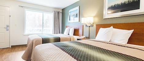 A hotel room with two beds, a large window, a painting on the wall, and a nightstand with a lamp.