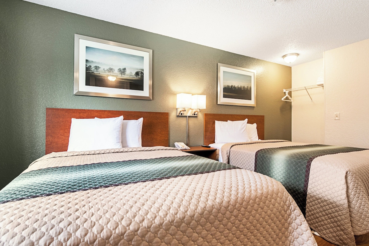 A hotel room with two beds, a nightstand, a lamp, and two framed pictures on the wall.