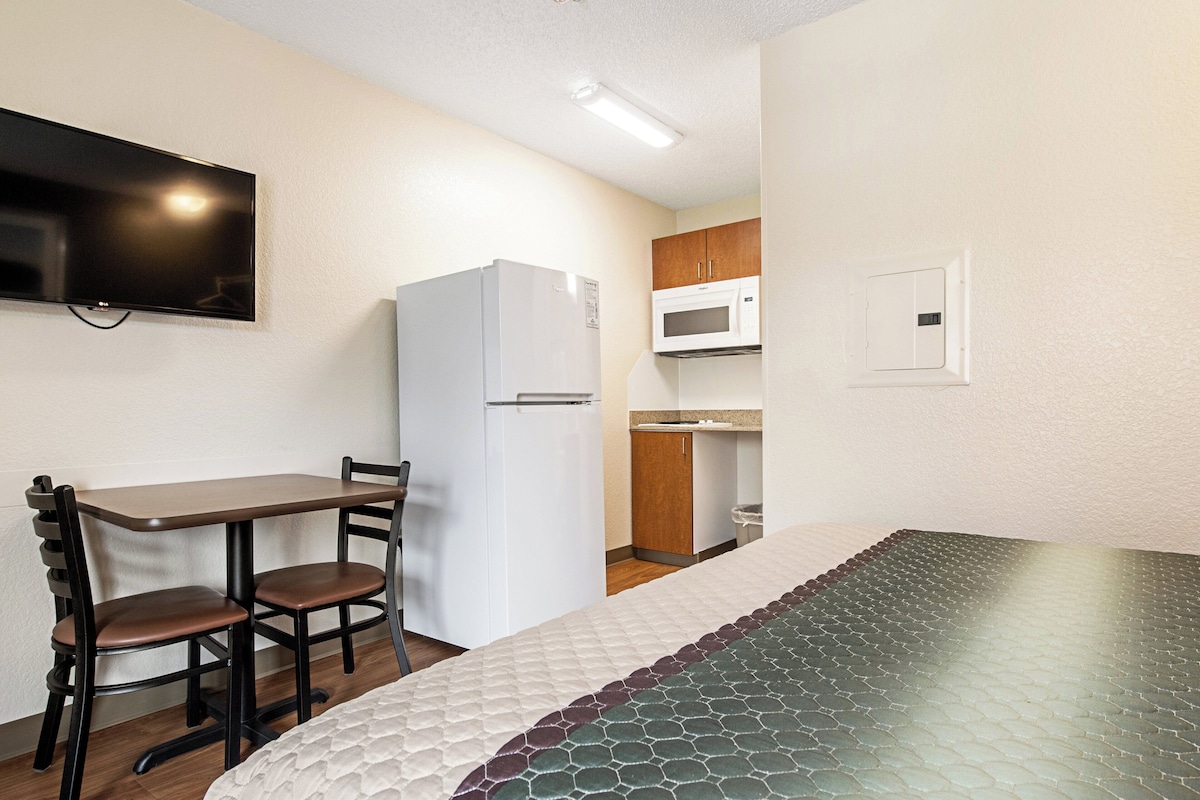 A compact room with a bed, a small table with chairs, a white refrigerator, a microwave, and a television mounted on the wall.