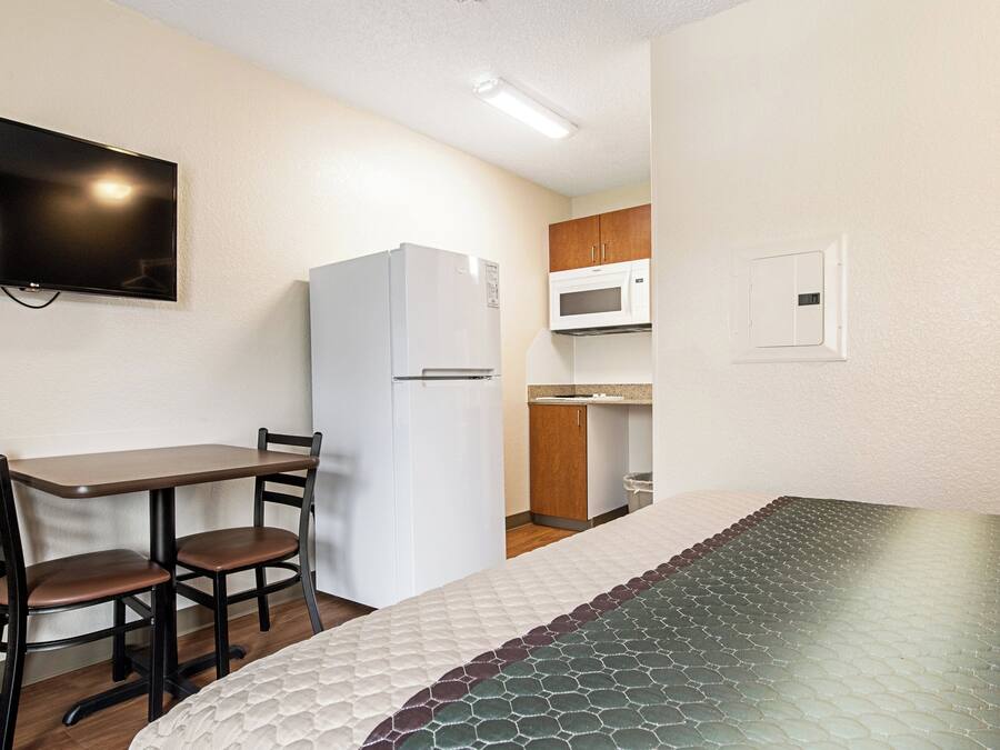 A compact room with a bed, a small table with chairs, a white refrigerator, a microwave, and a television mounted on the wall.